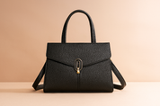 Structured black textured leather handbag with top handles, gold-tone hardware, and detachable shoulder strap, styled on a sandy pastel pink-lit background.