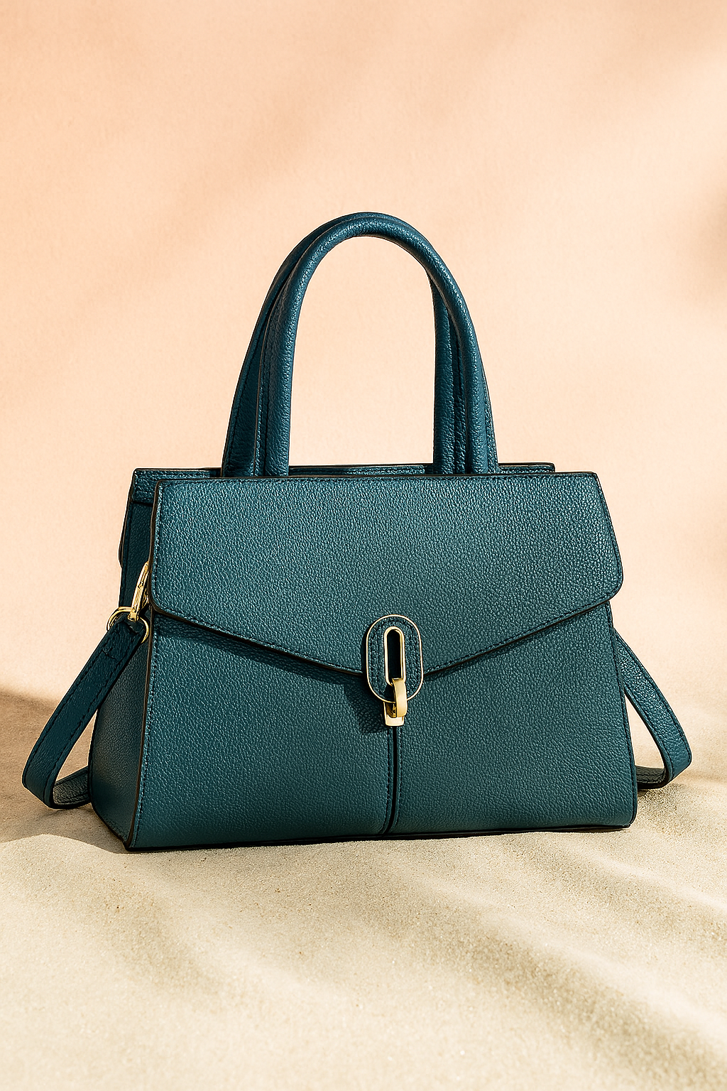 Teal pebbled leather handbag with gold-tone clasp, structured top handles, and detachable shoulder strap, shown on a soft sand base and warm pastel background.