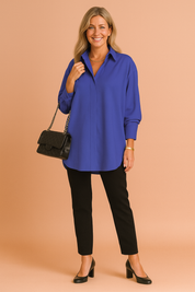 Royal blue oversized long-sleeve blouse with a clean collar and concealed front placket, paired with slim black trousers and classic black block-heeled pumps; accessorized with a quilted black handbag.