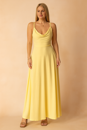 A yellow A-line maxi dress featuring a soft draped neckline and thin spaghetti straps, styled on a warm pastel background for a bright, elegant presentation.