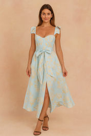 A sophisticated long summer dress in white with a sweetheart neckline, bow-tied waist, and ruffled shoulder straps, posing against a softly lit warm sand and pastel pink backdrop in a fashion shoot style.
