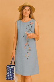 A woman wearing a sleeveless light blue summer dress with delicate pink floral embroidery cascading diagonally across the front. 