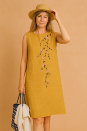 A woman wearing a sleeveless yellow summer dress with delicate pink floral embroidery cascading diagonally across the front. 