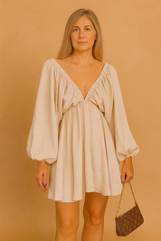 A beige mini dress with bishop sleeves, posing against a soft sand and pastel pink background in a fashion shoot style.