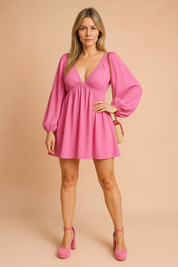 A pink mini dress with bishop sleeves, posing against a soft sand and pastel pink background in a fashion shoot style.