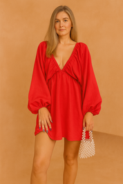A red mini dress with bishop sleeves, posing against a soft sand and pastel pink background in a fashion shoot style.