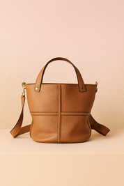 Medium-sized tan pebbled leather tote bag with structured cross-stitch detailing, gold-tone rivets, top handles, and a detachable shoulder strap, presented on a neutral display platform against a warm gradient background.