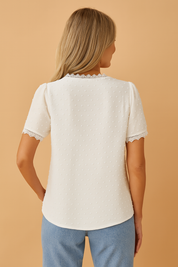 Elegant white short-sleeve blouse featuring delicate lace trim along the V-neckline and sleeves, textured dotted fabric, and a flattering straight cut, paired with light-wash high-waisted denim shorts and a woven beige belt.