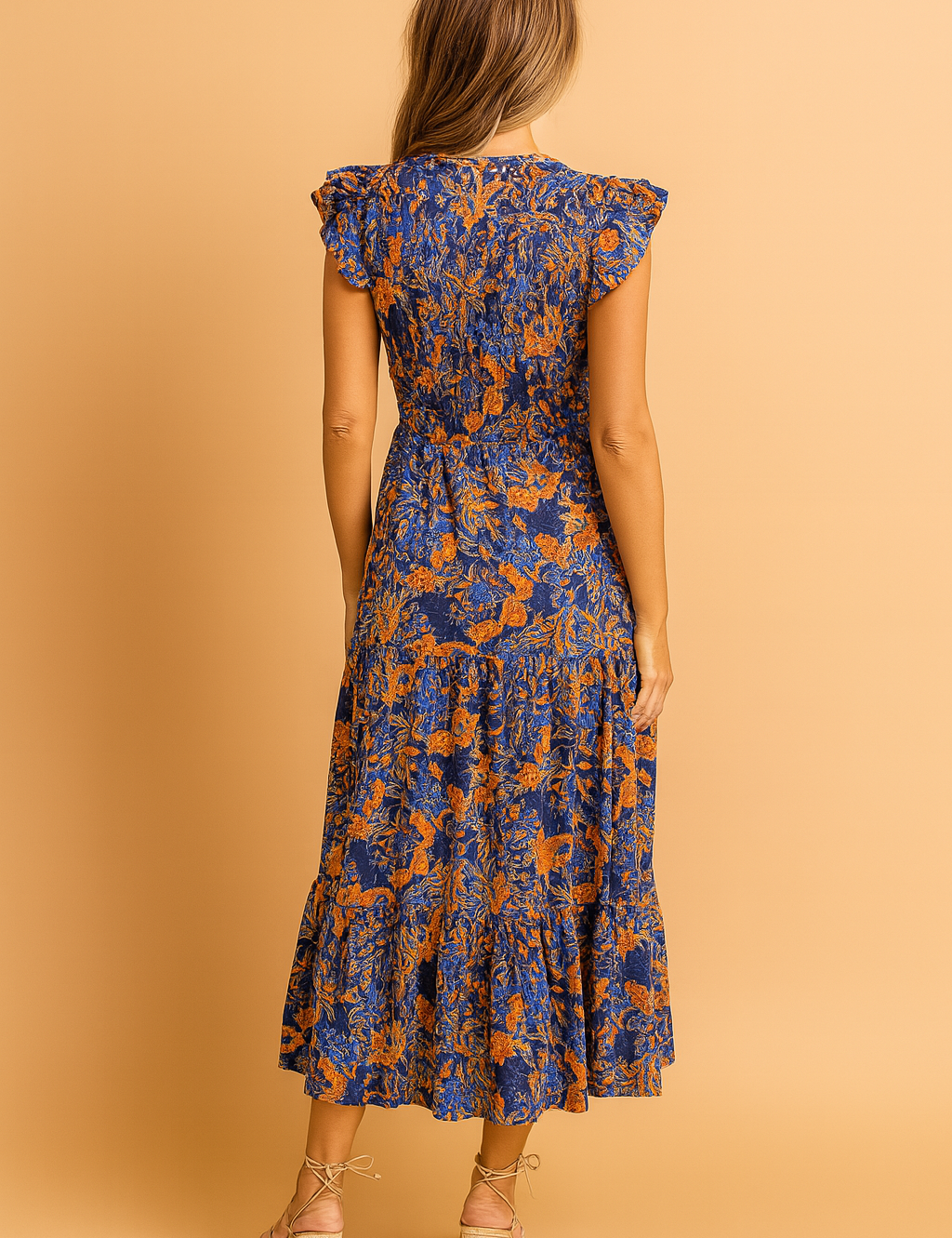 Back view of a navy blue and orange floral print midi dress with flutter sleeves, smocked bodice, defined waist, and a tiered hemline, featuring a lightweight flowy fabric with a vivid vintage-inspired botanical pattern.