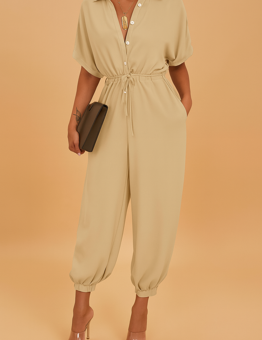 Beige jumpsuit with short sleeves, button-down front, and drawstring waist, finished with elasticized ankle cuffs, accessorized with a gold pendant necklace, transparent heels, and a sleek clutch.