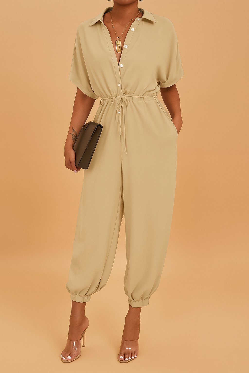 Beige jumpsuit with short sleeves, button-down front, and drawstring waist, finished with elasticized ankle cuffs, accessorized with a gold pendant necklace, transparent heels, and a sleek clutch.