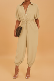 Beige jumpsuit with short sleeves, button-down front, and drawstring waist, finished with elasticized ankle cuffs, accessorized with a gold pendant necklace, transparent heels, and a sleek clutch.