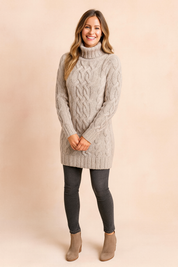 Beige cable-knit turtleneck sweater dress with long sleeves, paired with dark grey skinny jeans and taupe suede ankle boots.