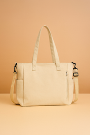 Beige canvas tote bag with dual top handles, side pockets, front zip compartment, and adjustable shoulder strap on a pastel backdrop.