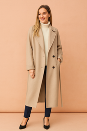 Beige long wool overcoat with a double-breasted button closure, wide lapel collar, side pockets, and a straight tailored cut, styled over a ribbed white turtleneck sweater and paired with navy slim-fit trousers and black pointed-toe heels.