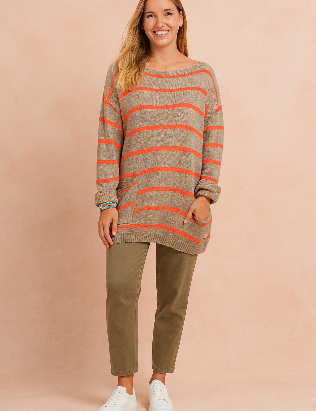 Beige oversized knit sweater with horizontal orange stripes, featuring a front pocket and rolled sleeves, paired with olive-green straight-leg pants and white low-top sneakers.