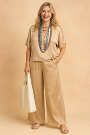 Beige satin co-ord set featuring a loose short-sleeve V-neck blouse and wide-leg high-waisted trousers, styled with layered beaded necklaces and paired with neutral-toned open-toe sandals and a soft structured cream tote bag.