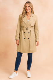 Classic beige trench coat with double-breasted black buttons, belted waist, wide lapels, and adjustable sleeve straps, paired with dark blue skinny jeans and white low-top sneakers.