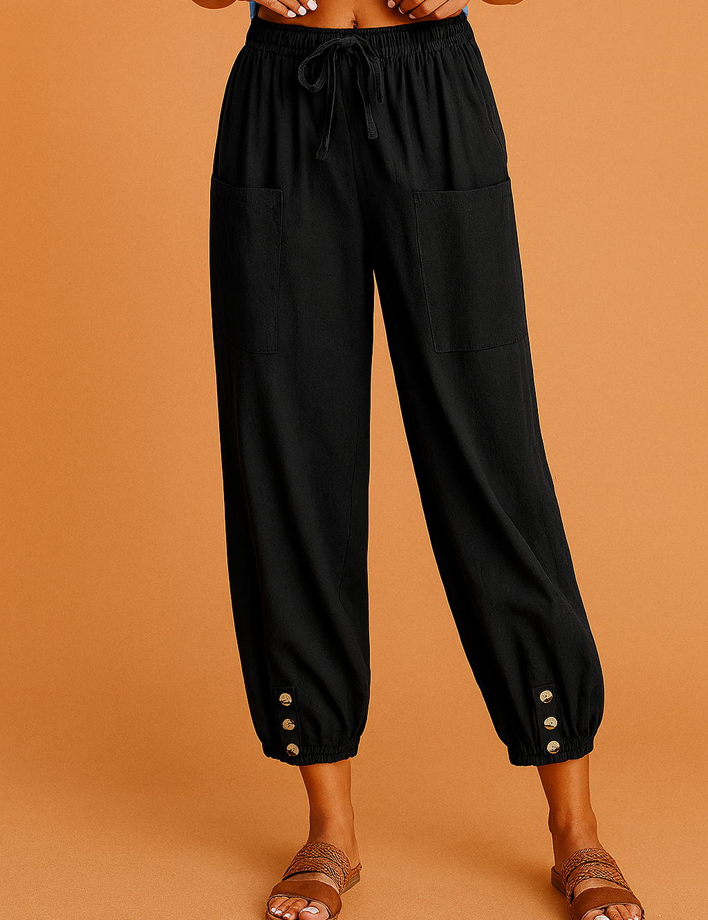 Black cropped drawstring pants with front patch pockets and gold-tone buttoned cuffs, styled with a blue knotted top and brown braided sandals on a warm sandy orange background.