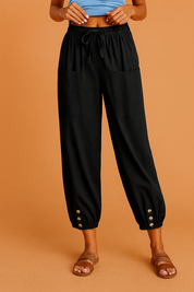 Black cropped drawstring pants with front patch pockets and gold-tone buttoned cuffs, styled with a blue knotted top and brown braided sandals on a warm sandy orange background.