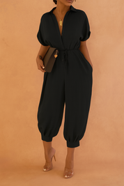 Black button-up jumpsuit featuring a drawstring waist, V-neck collar, and wide cropped legs with cuffed hems, paired with nude high heels and accessorized with a gold pendant and a sleek clutch.