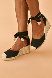 Black espadrille wedge sandals with natural jute soles, canvas uppers, cream-colored toe caps, and wraparound ankle ties with stitched square tips.