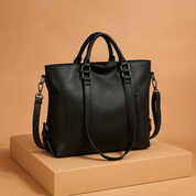 Black faux leather tote bag with grain texture, silver hardware, dual handles, and detachable strap, posed on a beige platform with a warm sand-to-pastel blush gradient background.
