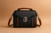 Elegant structured black handbag with brown accents, silver clasp, and textured finish, displayed on a soft sand-pink gradient background with gentle studio lighting.