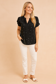 Short-sleeve black blouse with tiny white heart prints, pleated front detailing, and relaxed hem, paired with slim white trousers and white sandals.
