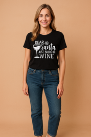 Black short-sleeve graphic t-shirt featuring bold white festive typography that reads "Dear Santa Just Bring Wine" with a wine glass illustration and snowflake accents, perfect for casual holiday wear.