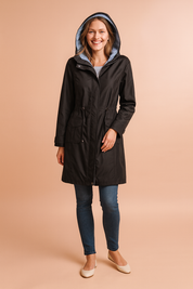 Black hooded raincoat with striped inner lining, front zipper closure, adjustable waist drawstring, flap pockets, and buttoned cuffs, styled with slim-fit blue jeans and cream flats.