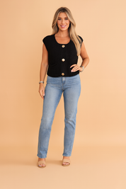 Black knit sleeveless button-front blouse with large gold buttons, styled with light-wash high-rise jeans and nude heeled sandals, photographed in a soft pastel fashion studio background