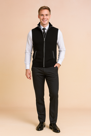 Black zip-up knitted vest with ribbed hem and pockets, layered over a crisp white dress shirt and paired with a black tie and tailored dark trousers.