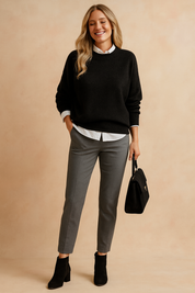 Black crewneck sweater layered over a white button-up shirt, paired with tailored grey trousers, black suede ankle boots, and a structured black handbag.