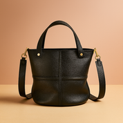 Black pebbled leather bucket bag with gold-tone hardware, structured seams, dual top handles, and detachable shoulder strap, displayed on a minimalist studio platform.