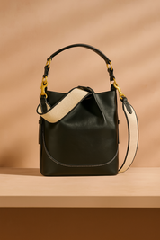 Black leather bucket bag with cream canvas strap, gold hardware, and contrast white stitching, displayed upright on a soft beige-pink gradient background.