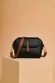 Black pebbled leather crossbody bag with brown strap and gold hardware, displayed on a beige pedestal against a warm gradient background.