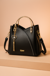 Black leather handbag with white contrast stitching, gold half-moon handles, detachable strap, and a dangling heart-and-pearl tassel charm, displayed on a beige platform with a soft pastel blush background.