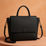 Modern black leather handbag with top handle, front flap closure, and detachable strap, displayed on a soft pink platform against a warm sand and pastel blush gradient background.