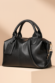 Black pebbled leather handbag with rounded handles, a side tag, and gold hardware, styled on a beige pedestal against a warm sand-to-blush gradient background.