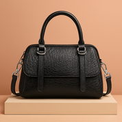 Black structured leather satchel handbag with textured finish, dual top handles, silver hardware, and detachable shoulder strap, styled on a studio platform.
