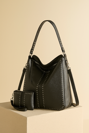 Black pebbled leather hobo bag with silver stud accents, adjustable shoulder strap, and matching studded zippered pouch, styled on a textured pedestal against a softly lit warm gradient backdrop.