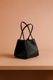 Structured black leather handbag with double top handles and pebbled finish on a pastel pink-beige gradient background.