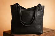 A black leather tote bag with front pocket, smooth finish, and double handles, displayed on a wooden platform.