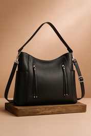 Black leather hobo handbag with dual top zippers and tassel accents, structured silhouette, and adjustable shoulder strap, displayed on a wooden base.
