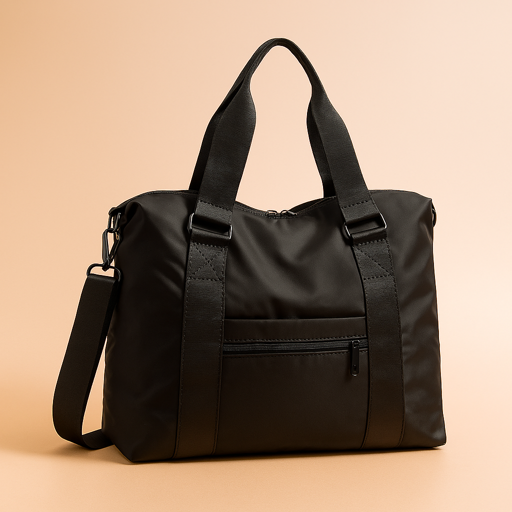 Black nylon shoulder bag with matte finish, wide webbing handles, front zip pocket, and adjustable strap, displayed on a warm sand-pink gradient background with soft studio lighting.