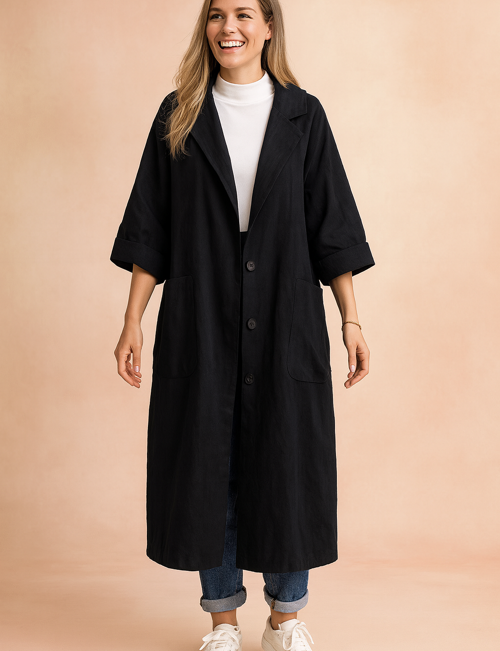 Long black oversized coat with wide sleeves, large front patch pockets, and button-down closure, styled with a white turtleneck, cuffed jeans, and white sneakers for a relaxed casual look.