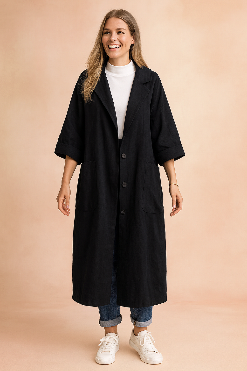 Long black oversized coat with wide sleeves, large front patch pockets, and button-down closure, styled with a white turtleneck, cuffed jeans, and white sneakers for a relaxed casual look.