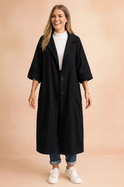 Long black oversized coat with wide sleeves, large front patch pockets, and button-down closure, styled with a white turtleneck, cuffed jeans, and white sneakers for a relaxed casual look.