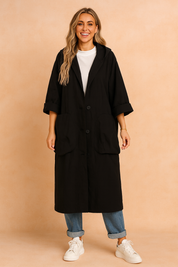Black oversized button-down coat with wide lapels, large front pockets, and rolled sleeves, styled over a white high-neck top, cuffed blue jeans, and white sneakers.